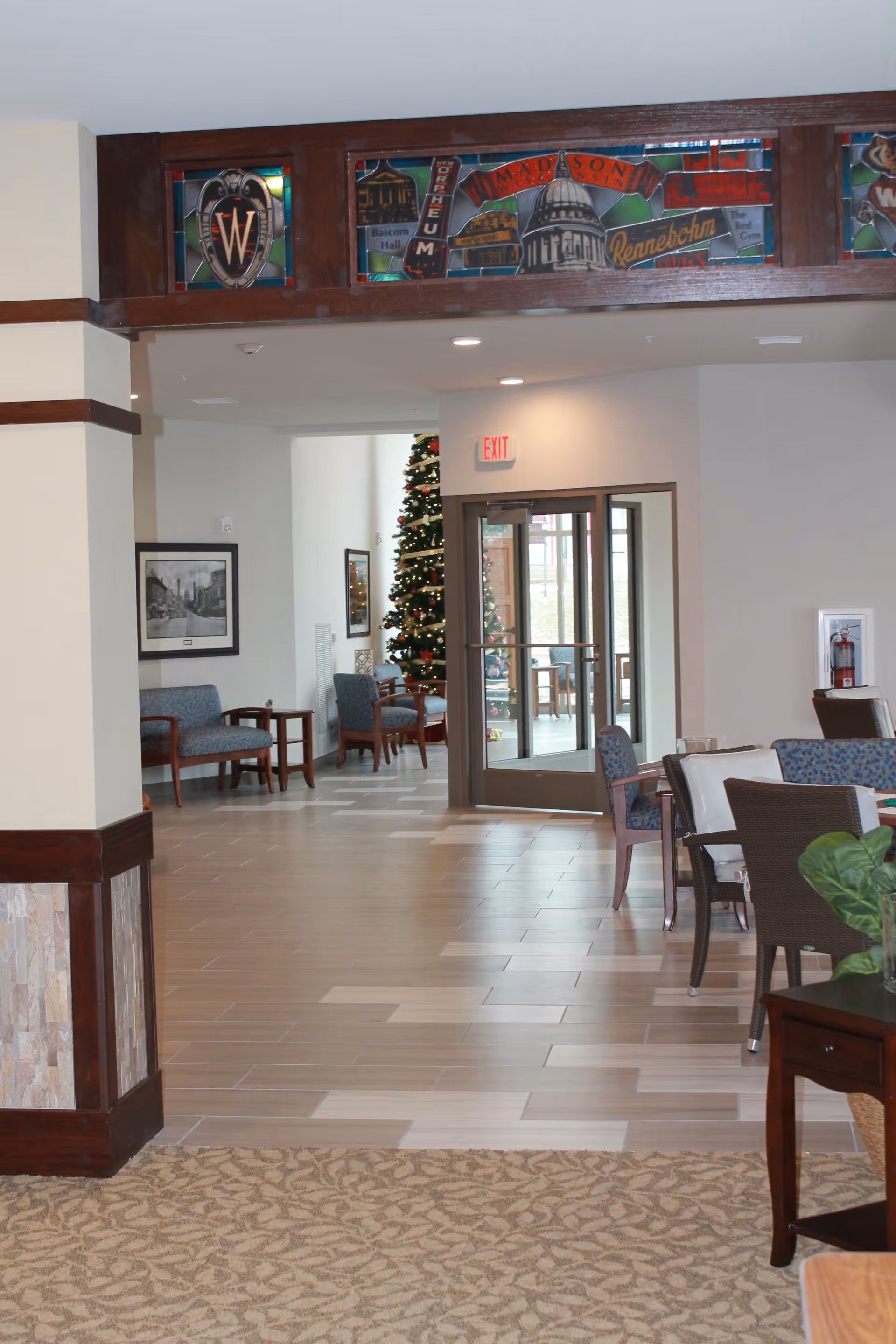 Interior view of a senior living facility lobby area with seating arrangements including chairs and small tables. A decorated Christmas tree is visible in the background near glass doors with an exit sign above. The floor transitions from carpet to tile, and there is decorative stained glass artwork above the entrance.