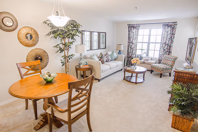 Bright living room with a beige sofa adorned with decorative pillows, a round wooden coffee table with a flower vase, a patterned armchair near a large window with floral curtains, a wooden dining table with two chairs, a tall potted plant, and wall decorations including round mirrors and framed pictures.