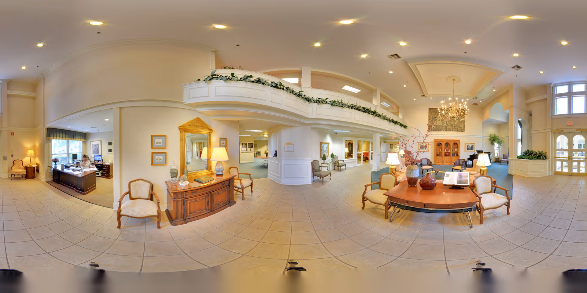 A spacious and well-lit senior living facility lobby with tiled floors, cream-colored walls, and high ceilings. The area features a wooden reception desk with a staff member seated, several upholstered chairs, a wooden console table with a large mirror above it, and a central round table decorated with vases and lamps. There is a chandelier hanging from the ceiling and large windows allowing natural light to enter. The space has a welcoming and elegant atmosphere with plants and framed artwork on the walls.