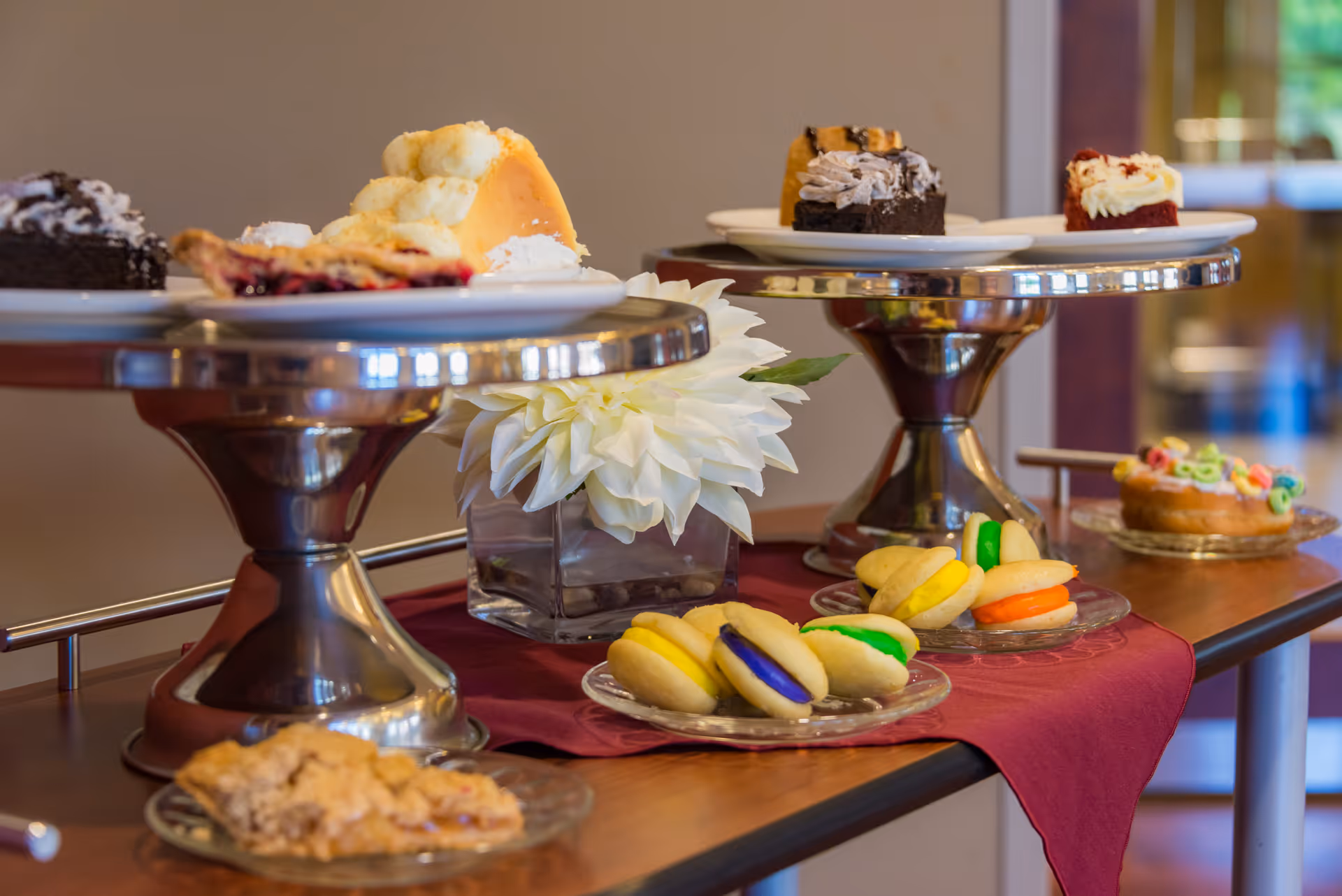 A dessert display on a wooden table with a burgundy cloth, featuring various sweet treats including colorful macarons, slices of cake, and cookies, with a white flower centerpiece in a glass vase.