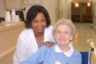 A smiling elderly woman in a wheelchair wearing glasses and a blue sweater is accompanied by a smiling female caregiver in a white coat standing behind her in a hallway of a nursing facility.
