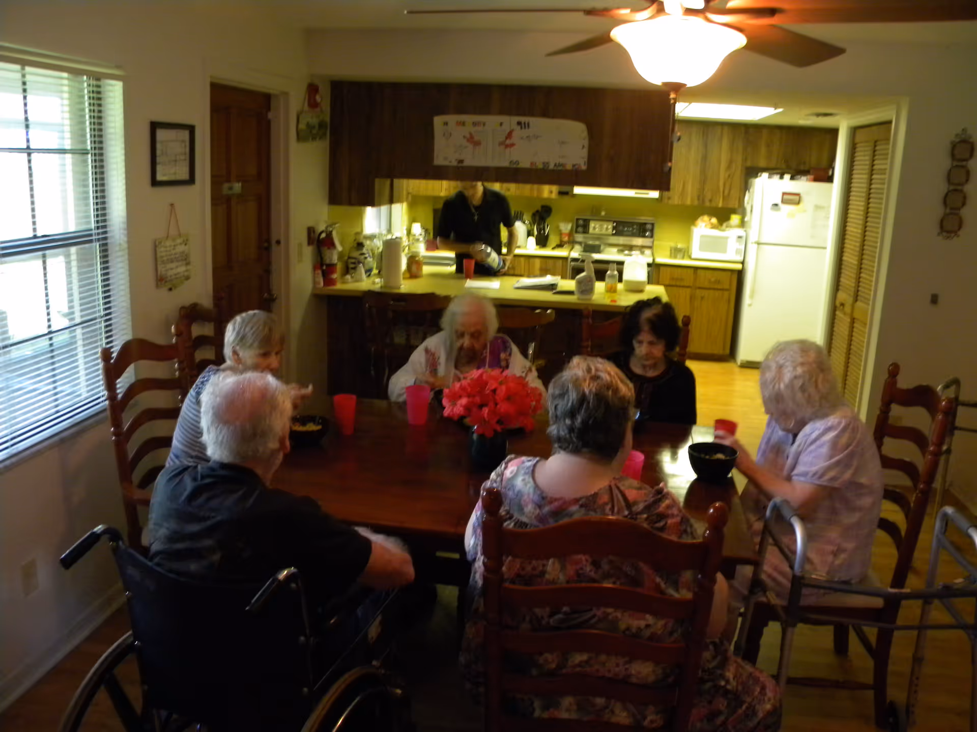 A group of elderly people sitting around a wooden dining table in a home-like setting, with a kitchen visible in the background. One person is in a wheelchair and another uses a walker. A person is standing in the kitchen area preparing or serving food. The room is warmly lit with a ceiling fan light above the table.