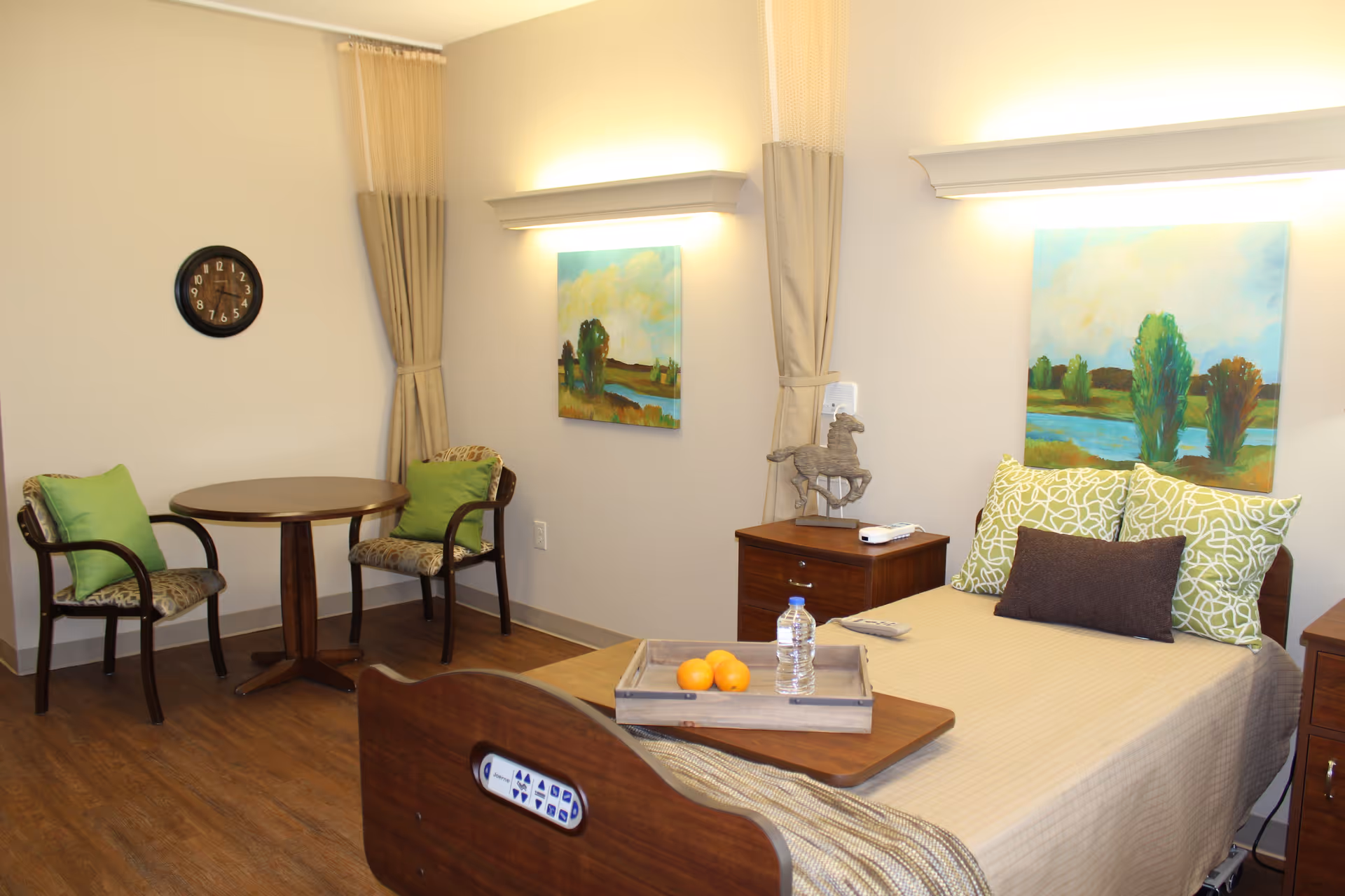 A furnished patient bedroom with a single bed, bedside tables, artwork on the walls, a tray with water and fruit, and a small round table with two chairs.