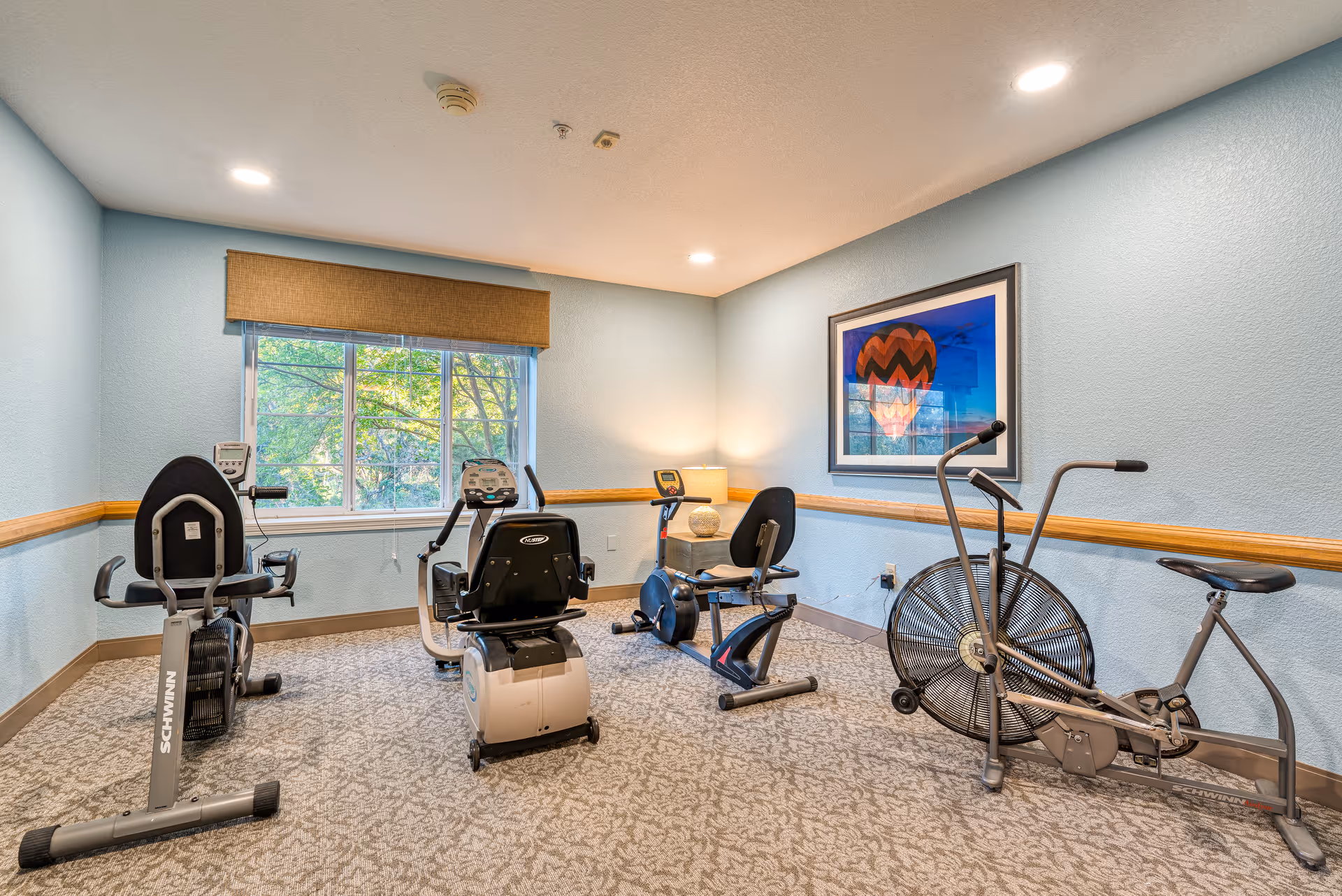 A small exercise room with light blue walls and beige patterned carpet. The room contains four pieces of exercise equipment, including two recumbent stationary bikes, one upright stationary bike, and one fan bike. There is a window with a brown valance letting in natural light, a framed picture of a hot air balloon on the wall, and a small table with a lamp in the corner.