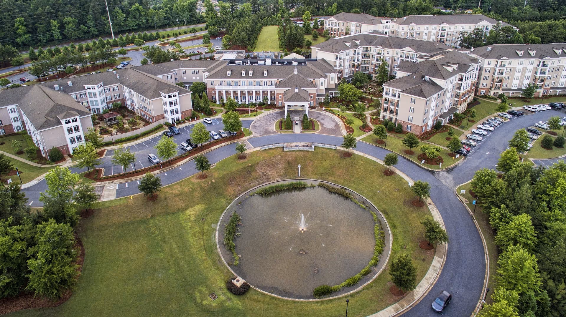 Aerial view of Spring Harbor at Green Island senior living facility showing multiple connected buildings surrounding a circular driveway with a central water fountain and landscaped greenery.