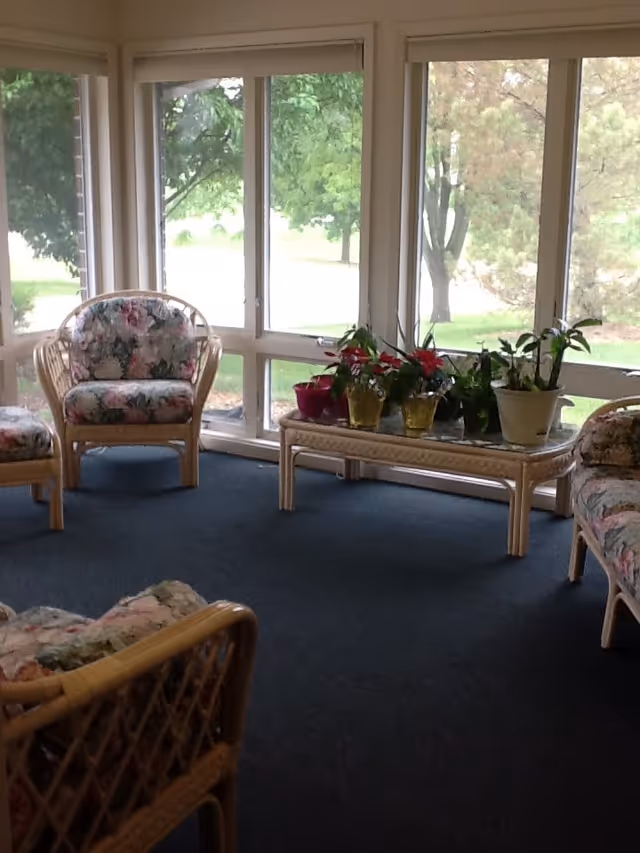 A sunlit sitting area with large windows overlooking green trees outside. The room has floral cushioned wicker chairs and a wicker coffee table with several potted plants on it. The floor is carpeted in dark blue.