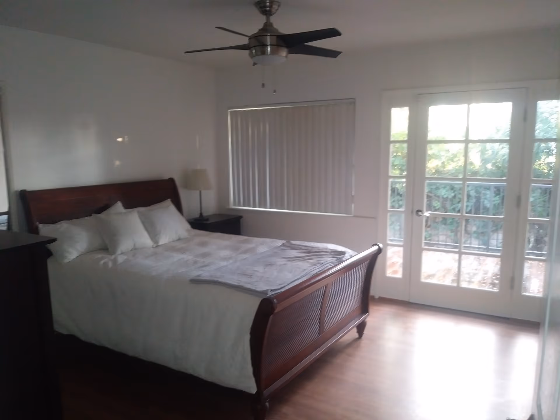 A bedroom with a wooden bed frame, white bedding, and several pillows. There is a nightstand with a lamp next to the bed, a ceiling fan above, a window with vertical blinds, and glass doors leading outside to a balcony or patio area with greenery visible.