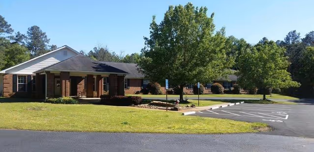 Single-story brick building with a covered entrance, surrounded by green grass, trees, and a parking lot with marked handicap spaces under a clear blue sky.