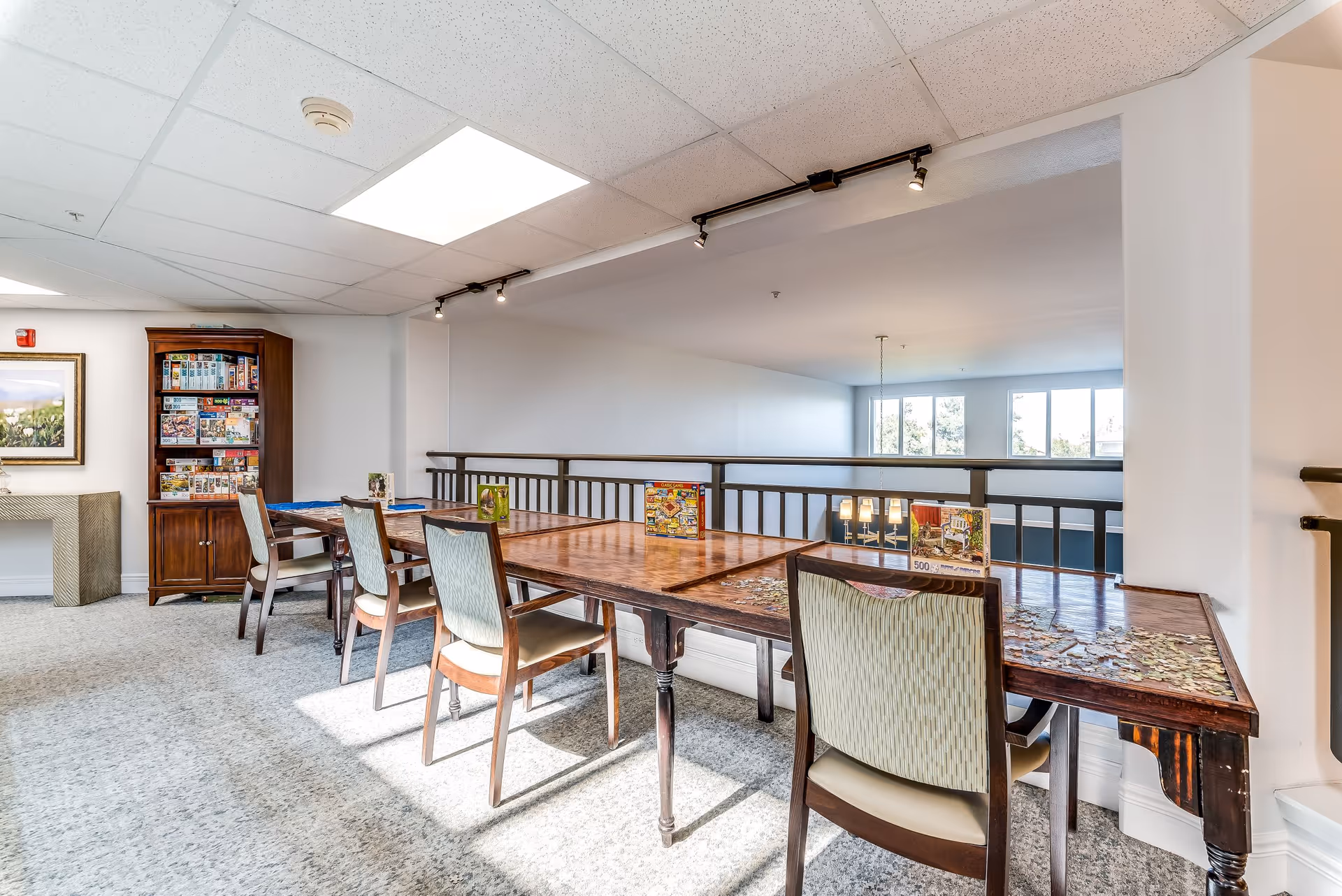 A bright room with a long wooden table and several chairs arranged around it. The table has multiple jigsaw puzzles in progress with puzzle boxes displayed on top. In the background, there is a wooden cabinet filled with more puzzle boxes and a framed picture on the wall. Large windows allow natural light to fill the space.