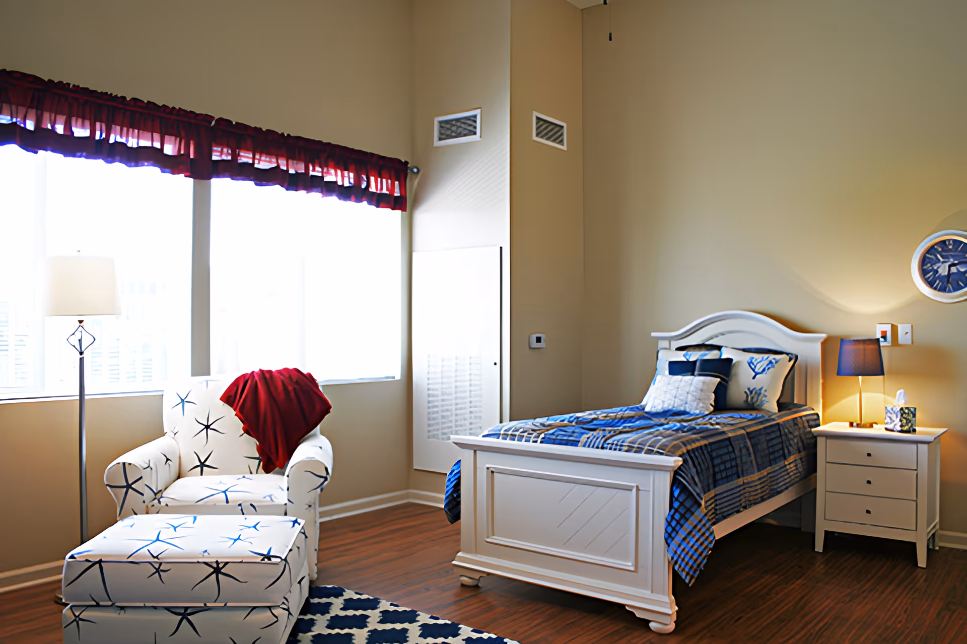 Well-lit bedroom with a single white bed, nightstand, and a patterned armchair by large windows.
