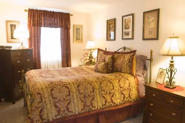 A warmly lit bedroom with a large bed covered in a gold and burgundy patterned bedspread and matching pillows. The room features two bedside tables with lamps, framed artwork on the walls, a window with burgundy curtains, and a dark wooden dresser.