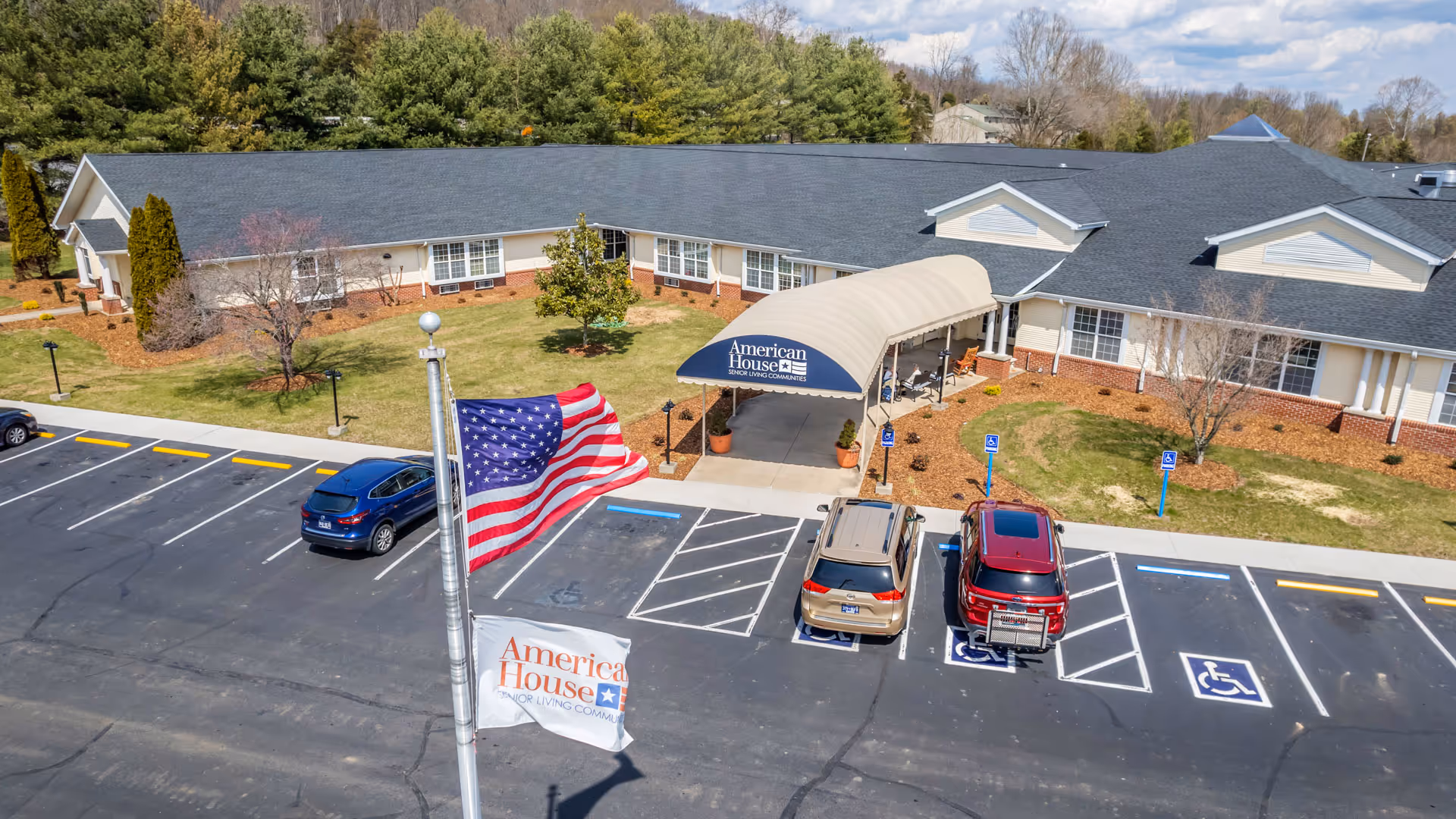 Exterior view of American House Johnson City senior living community building with a covered entrance, parking lot with handicap spaces, and two flags on a flagpole in the foreground. Trees and a partly cloudy sky are visible in the background.
