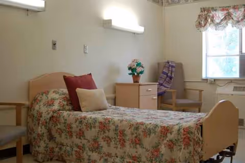Single floral bed with pillows, nightstand, plant, chairs, and window in a senior living bedroom.