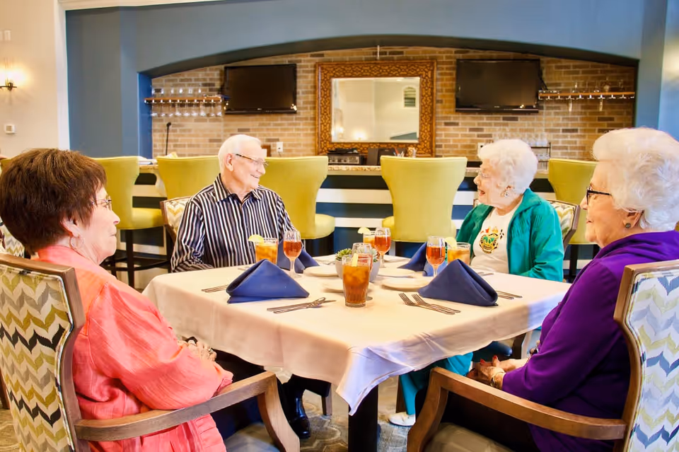 Four elderly people sitting around a dining table in a well-lit room, engaging in conversation. The table is set with blue napkins, glasses of iced tea, and silverware. The background features a brick wall with two mounted televisions and a large decorative mirror between them, along with green upholstered chairs at a counter.