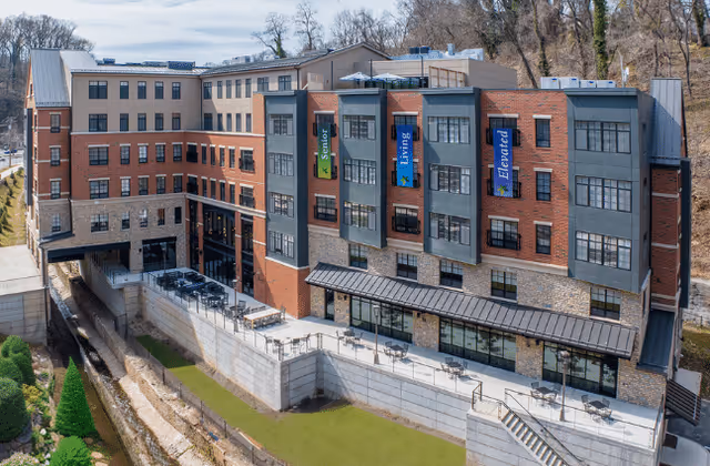 Exterior view of a multi-story senior living facility building with a combination of brick and stone facade. The building has large windows and three vertical banners that read 'Senior', 'Living', and 'Elevated'. There is an outdoor terrace area with tables and chairs overlooking a small grassy area and a creek. Trees and a wooded area are visible in the background.