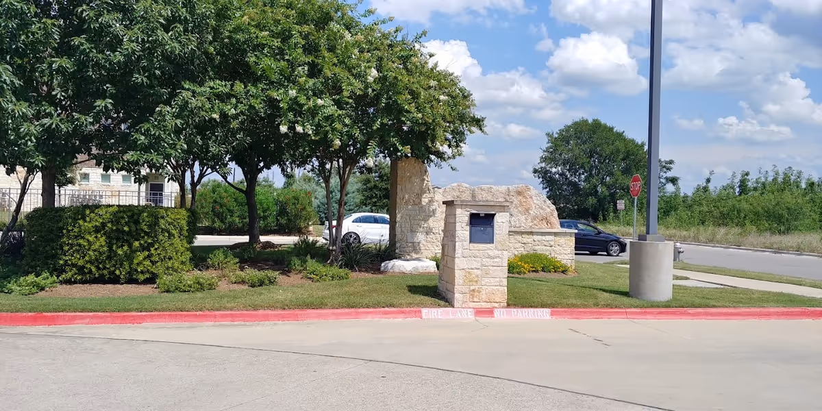 Outdoor view of a landscaped area with trees, bushes, and a stone mailbox structure near a street curb painted red with 'FIRE LANE NO PARKING' written on it. A stop sign and a streetlight are visible in the background under a partly cloudy sky.