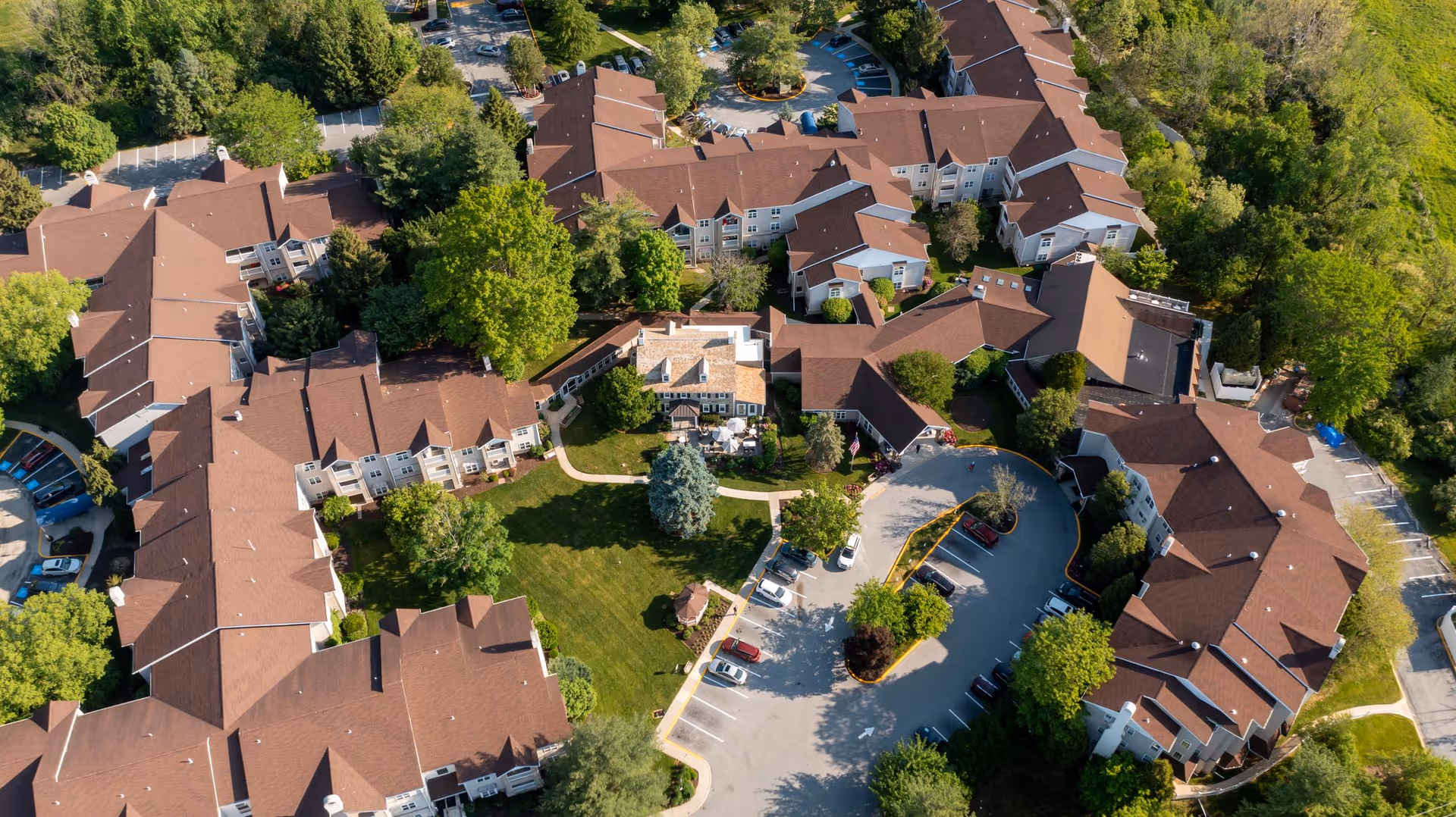 Aerial view of Bellingham at West Chester senior living facility showing multiple connected buildings with brown roofs surrounding a central green lawn area with trees and pathways, adjacent parking lots with cars, and lush greenery around the property.