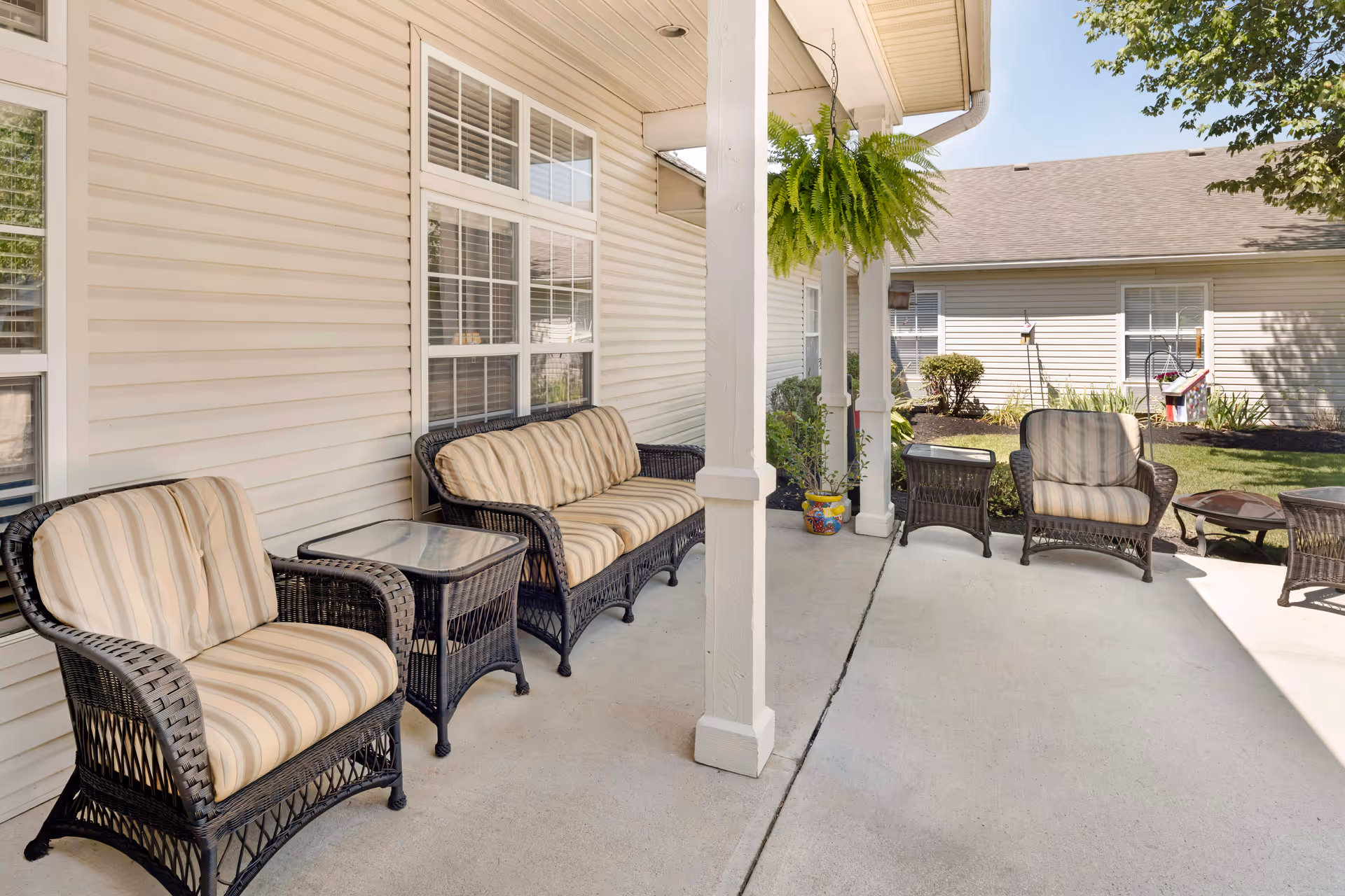 Outdoor patio area at Brookdale Piqua with wicker furniture including two armchairs, a loveseat with striped cushions, glass-top side tables, a hanging fern plant, and a fire pit on a concrete floor. The patio is adjacent to a beige building with white-trimmed windows and surrounded by a garden with bushes and a tree.