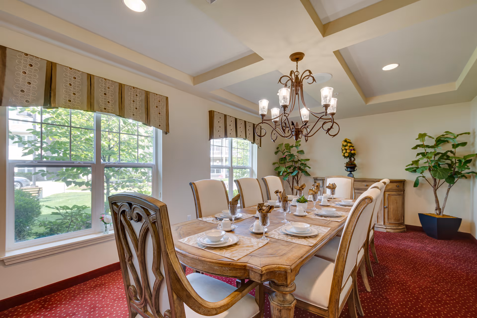 A well-lit dining room with a wooden dining table set for eight people. The table has beige placemats, white plates, bowls, cups, and glasses with brown napkins. The room features large windows with patterned valances, a decorative chandelier hanging from a coffered ceiling, red carpet flooring, and two large potted plants in the corners.