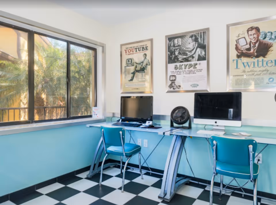 Small computer room with two desktop computers on a wall-mounted counter, teal chairs, vintage social media posters, a large window with palm-tree view, and a black-and-white checkered floor.
