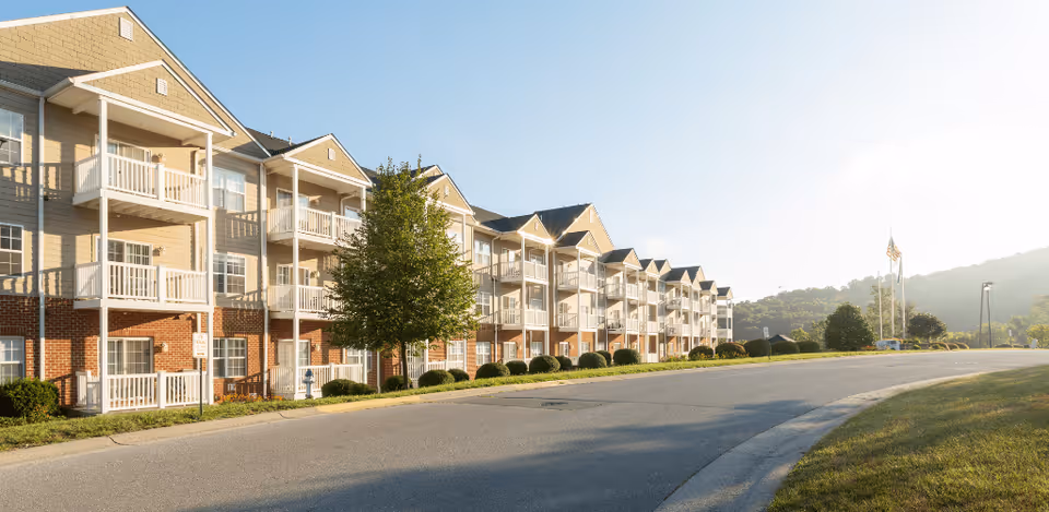 Exterior view of a multi-story senior living facility building with balconies, a tree, and a curved driveway under a clear sky with hills in the background.