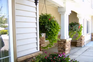 Covered front porch with white siding, stone-column bases, hanging flower baskets, and planted flowers along a sidewalk.