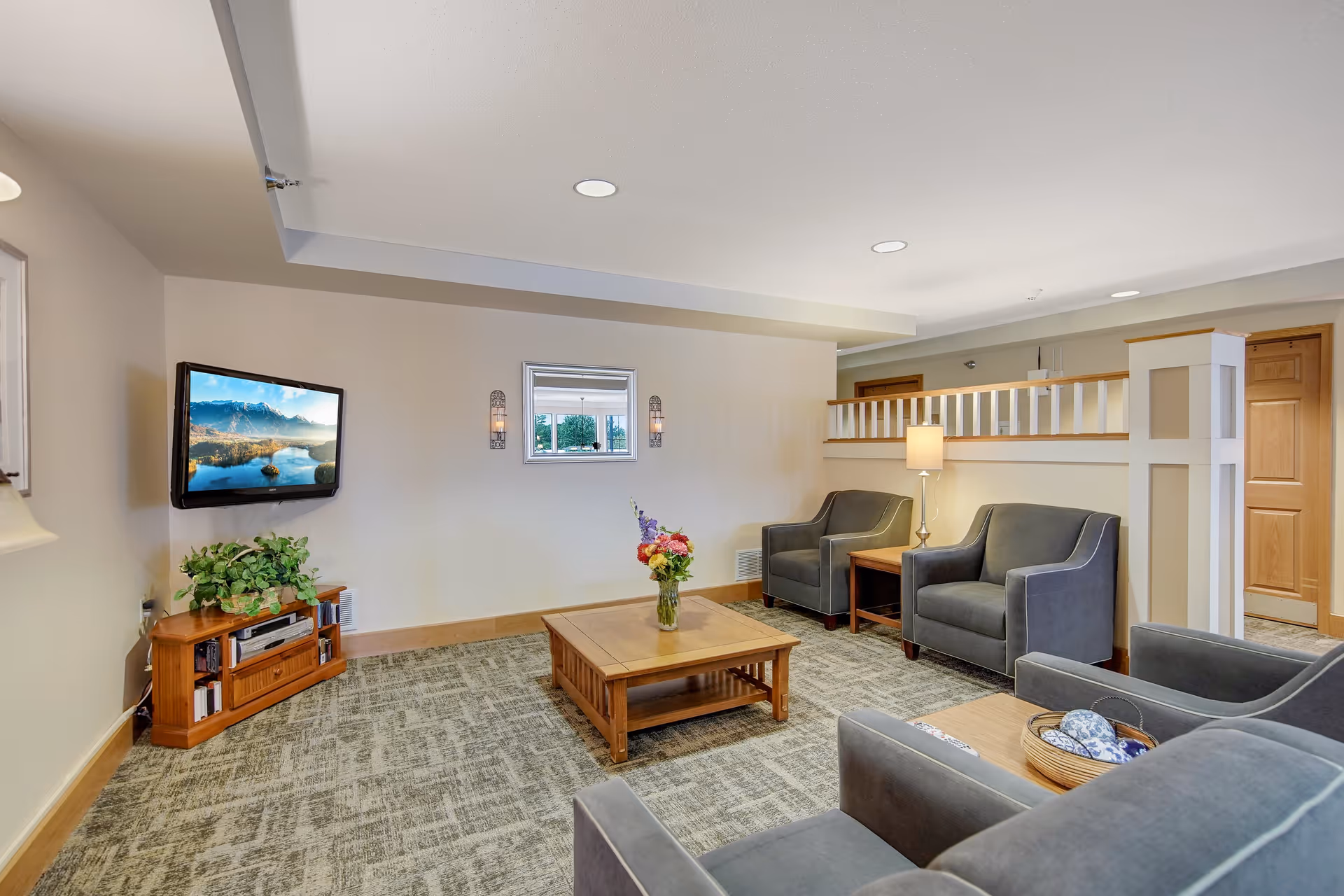A cozy living room area with gray upholstered armchairs and a matching sofa arranged around a wooden coffee table with a vase of flowers. A flat-screen TV is mounted on the wall above a wooden media console with a plant and electronic devices. The room has beige walls, carpeted flooring, recessed lighting, and a small window with two wall sconces on either side.