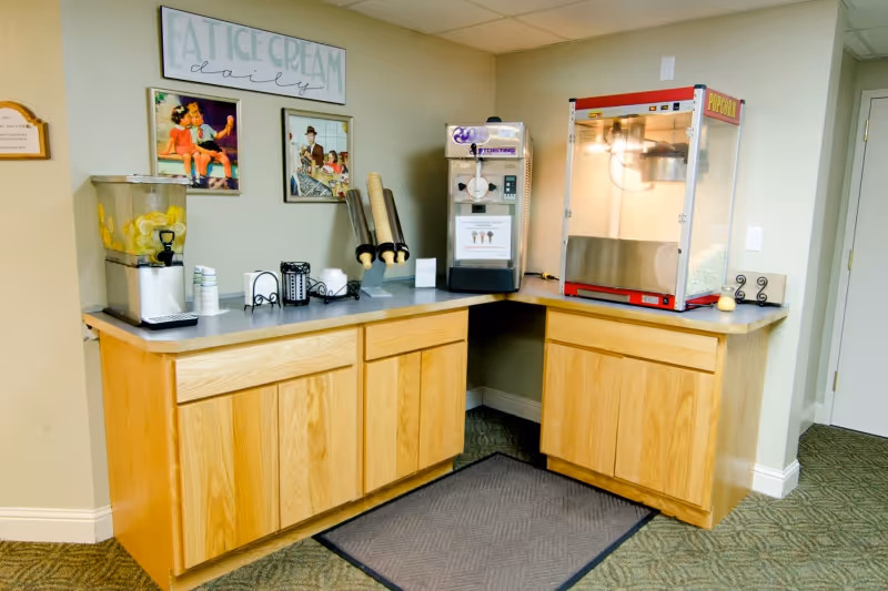 A snack and beverage corner in a facility with wooden cabinets and a gray countertop. On the counter, there is a dispenser with lemon water, a napkin holder, a soft serve ice cream machine, and a popcorn machine. Above the counter, there are three framed pictures and a sign that reads 'EAT ICE CREAM daily'. The floor has a carpet with a small rug in front of the cabinets.