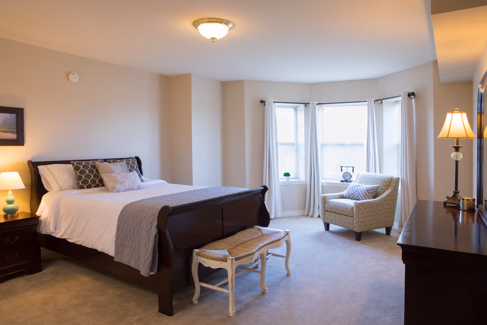 A spacious bedroom with a large wooden bed made with white and gray bedding, a cushioned bench at the foot of the bed, a nightstand with a lamp on the left, a comfortable armchair near a bay window with white curtains, and a wooden dresser with a lamp and mirror on the right.