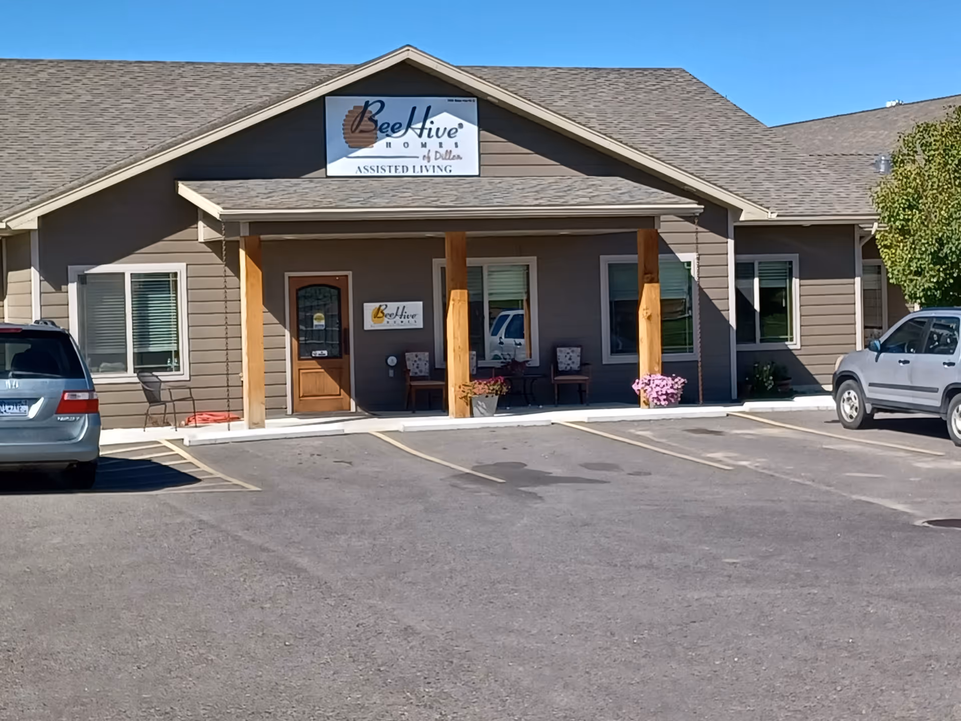 Front exterior view of BeeHive Homes of Dillon assisted living facility with a parking lot in front, two cars parked, a covered entrance supported by wooden posts, and a sign above the entrance displaying the facility's name.