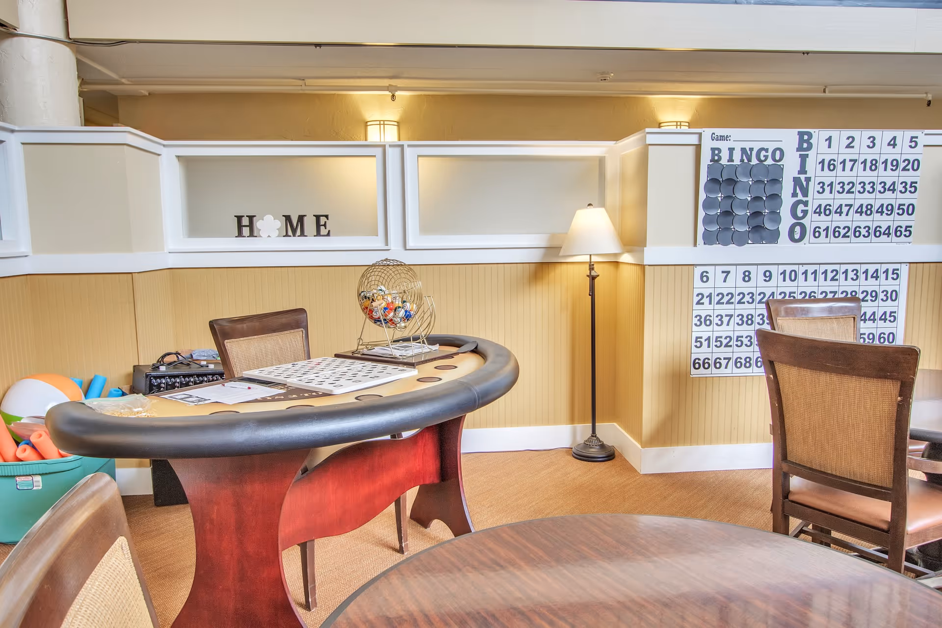 A cozy game room with a bingo table and chairs. The table has bingo cards and a bingo ball cage on it. On the wall, there are large bingo number boards and a decorative sign that spells 'HOME'. A floor lamp provides warm lighting in the corner.