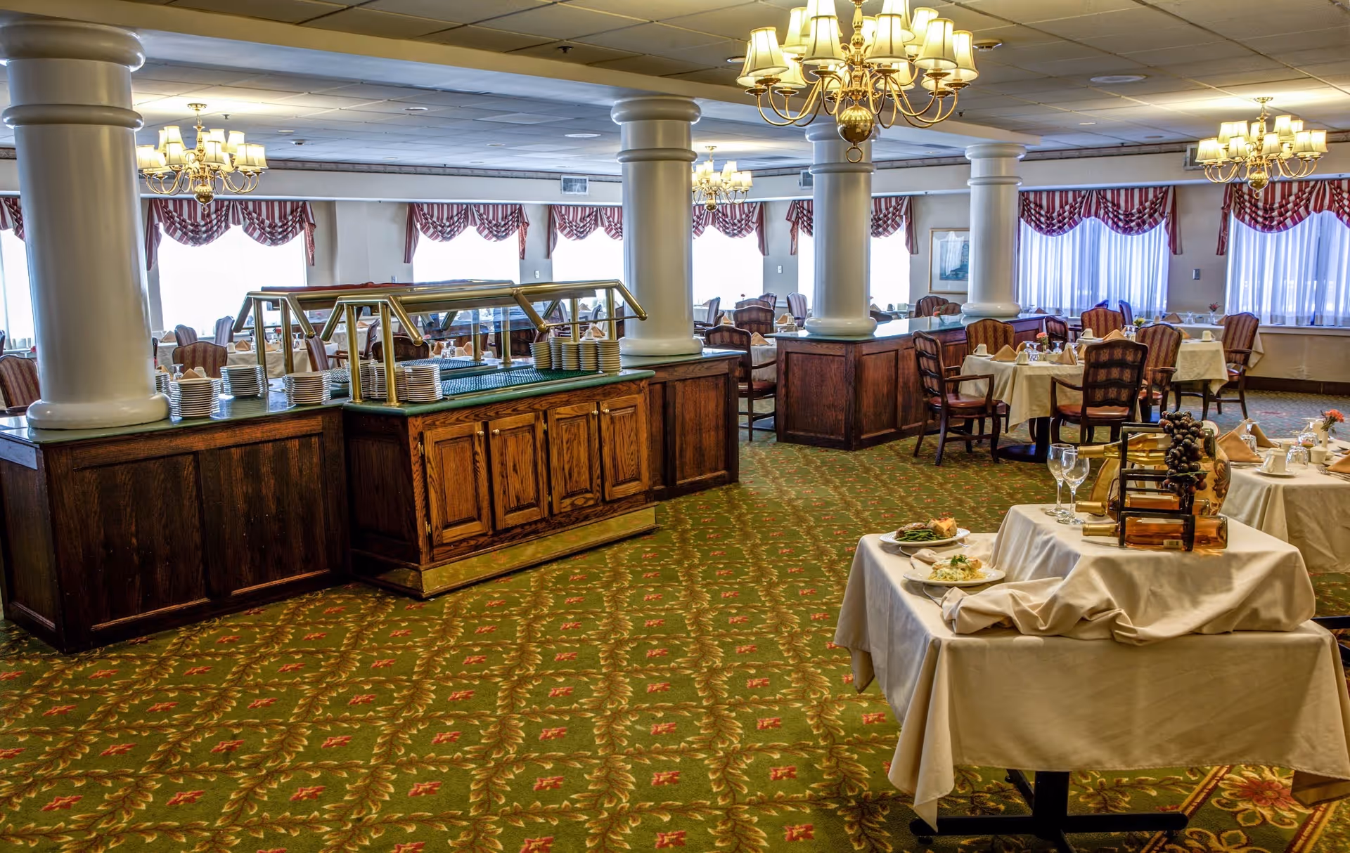 A spacious dining room with multiple round tables covered with white tablecloths, set with plates, glasses, and silverware. There are wooden chairs around the tables and a buffet counter with stacks of plates. The room has large windows with red and white valances, green patterned carpet, white pillars, and chandeliers hanging from the ceiling.