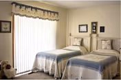 Sunlit bedroom with two twin beds dressed in blue and cream quilts beside a sliding glass door with vertical blinds.
