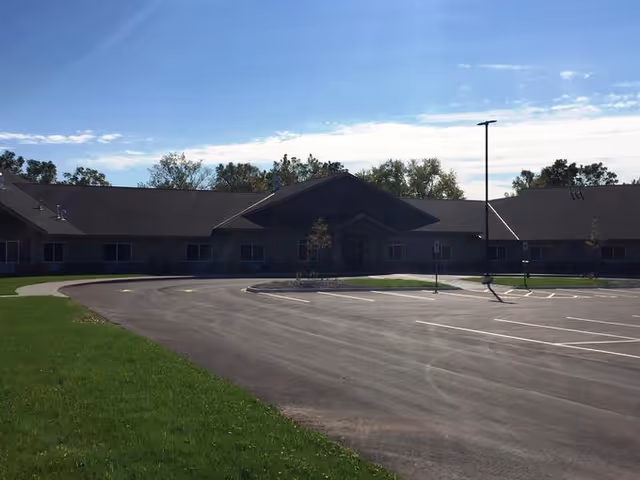 Exterior view of a single-story assisted living facility building with a large paved parking lot in front, surrounded by green grass and trees under a partly cloudy blue sky.