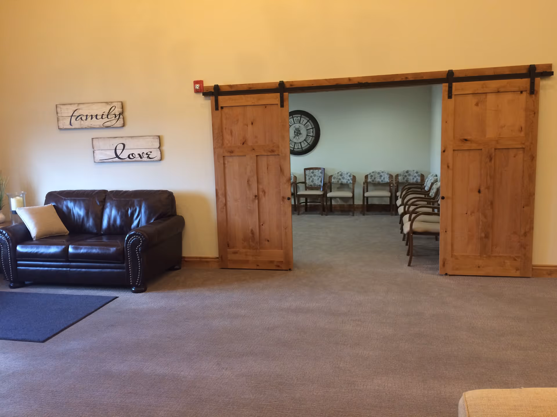 Interior view of a living area with a dark brown leather loveseat on the left, a beige pillow on it, and two wooden sliding barn doors leading to a room with several chairs arranged along the walls and a large clock on the far wall. Two decorative wooden signs on the wall say 'family' and 'love'.