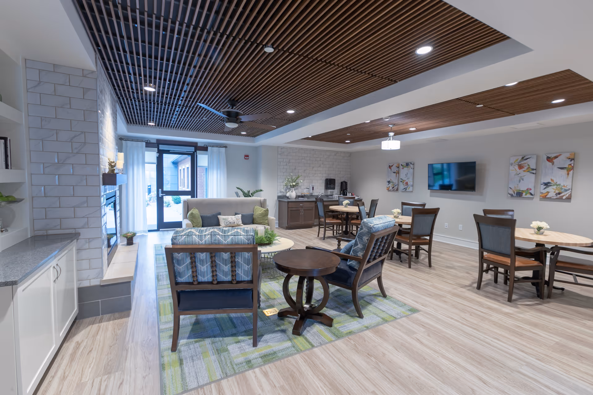 A bright and spacious common area in a senior living facility featuring comfortable seating with armchairs and a sofa around a coffee table on a patterned rug. There are several round dining tables with chairs, a wall-mounted flat screen TV, and decorative artwork on the walls. The ceiling has wooden slats with recessed lighting and ceiling fans. Large windows and a glass door allow natural light to enter the room.
