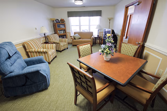 A cozy living room with a blue recliner, two striped armchairs, a beige loveseat, a wooden bookshelf, a TV on a stand, and a wooden table with four chairs. A vase with flowers is placed on the table. The room has a window with a valance, two floor lamps, and a door slightly ajar.