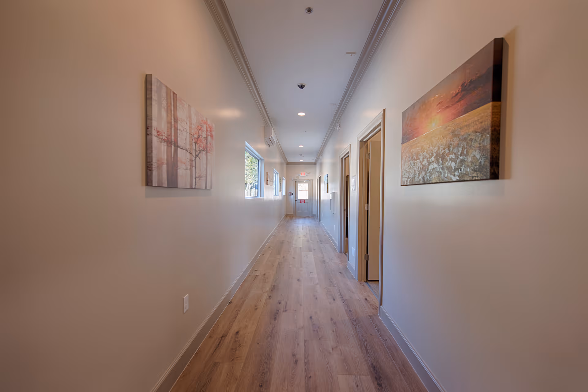 A long, narrow hallway with light-colored walls and wood flooring. There are several doors on the right side and two paintings hanging on the walls. A window on the left side lets in natural light, and the hallway leads to a door at the far end.