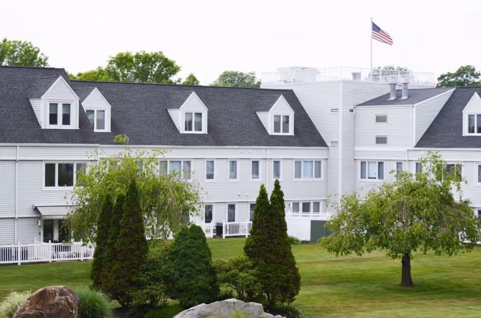Exterior view of a senior living facility building with white siding and dark gray roof. The building has multiple windows and dormers, with a well-maintained lawn, trees, and shrubs in the foreground. An American flag is flying on a flagpole on the roof.