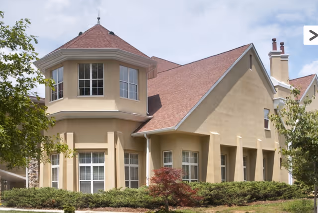 Exterior view of a beige two-story building with a red roof, multiple windows, and surrounded by green shrubs and trees under a partly cloudy sky.