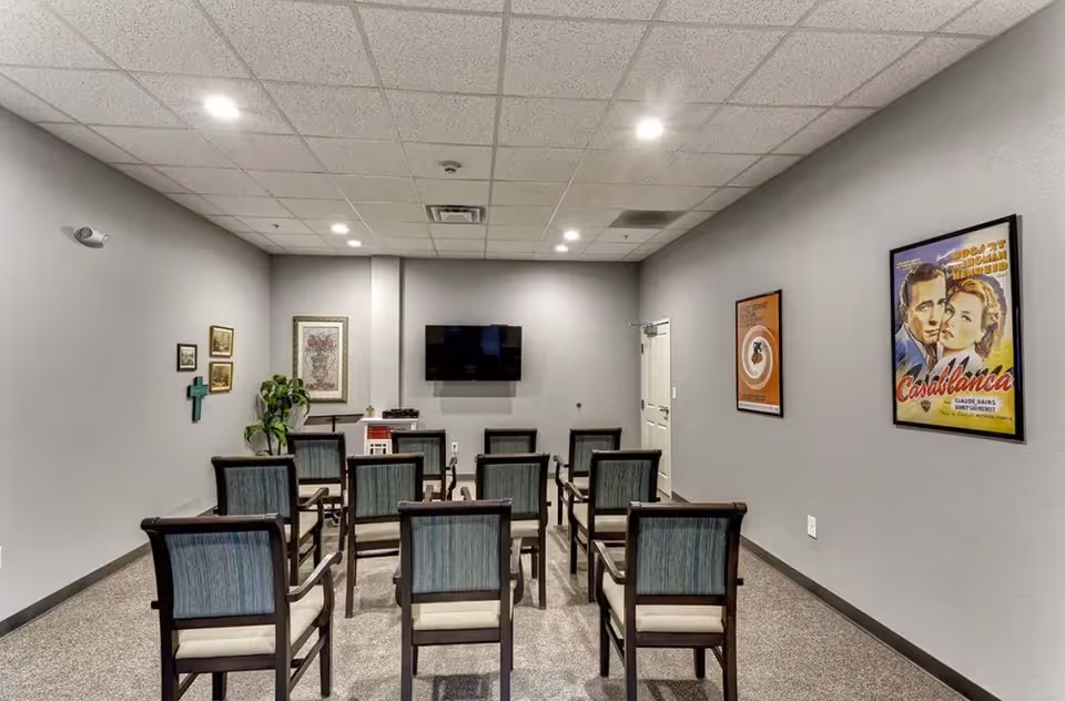 Small meeting/media room with rows of chairs facing a wall-mounted TV and framed posters on gray walls.