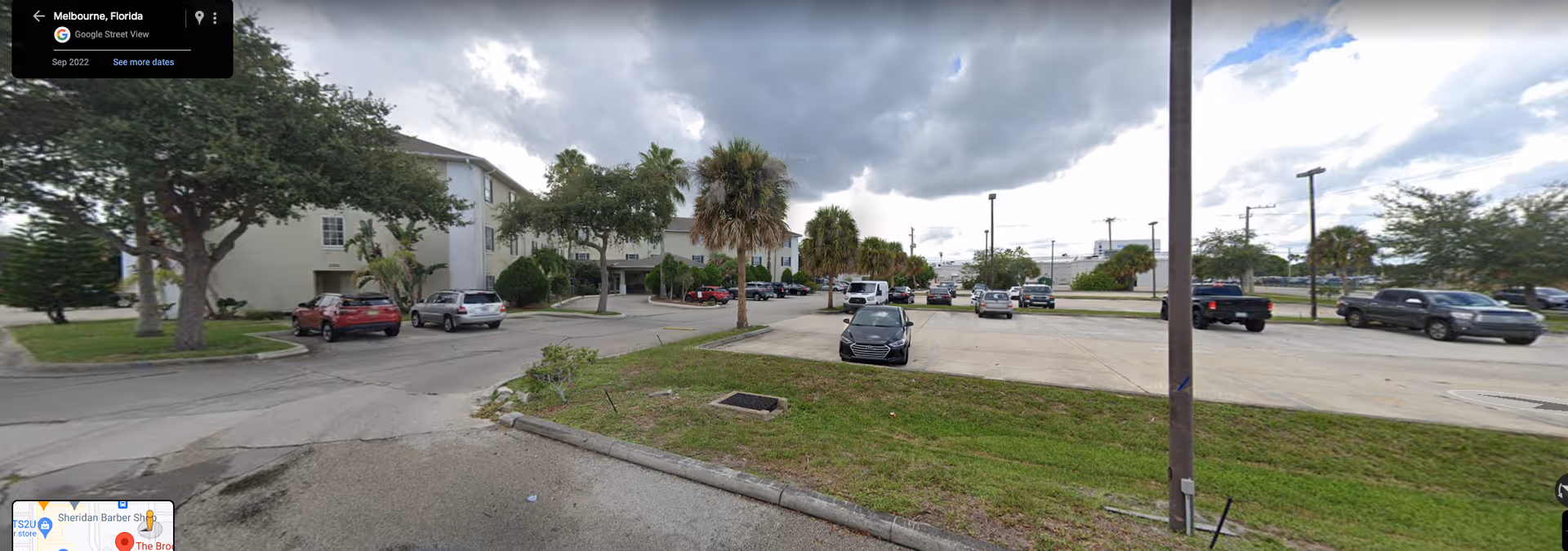 Parking lot with several cars parked near a multi-story beige building surrounded by trees and palm trees under a cloudy sky in Melbourne, Florida.