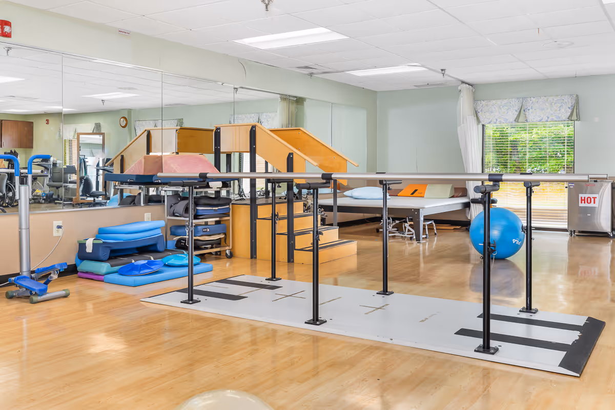 Physical therapy room with parallel bars, therapy stairs, treatment tables, exercise ball and stacked mats.