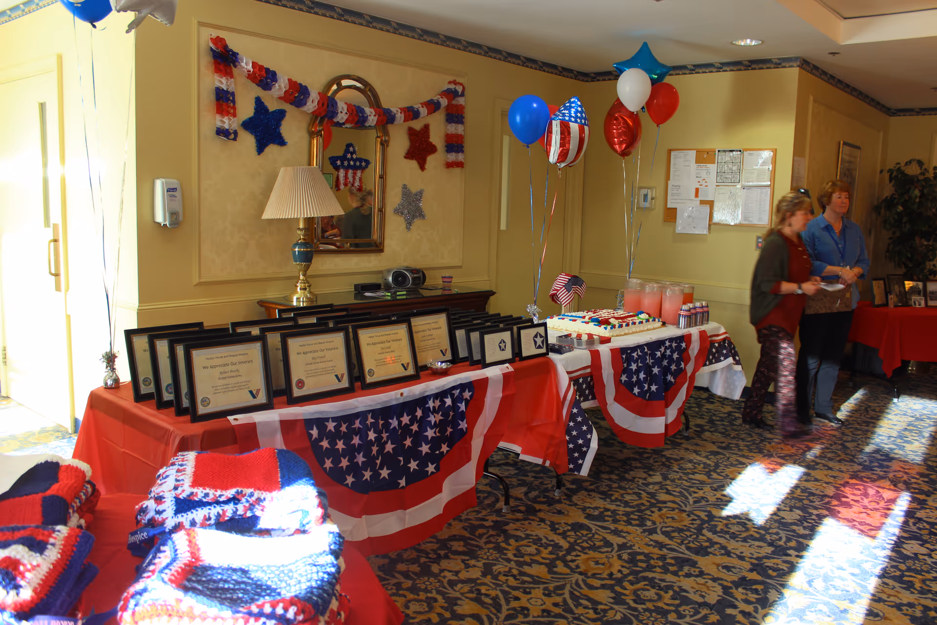 A patriotic-themed reception table in a facility lobby displaying framed certificates, a sheet cake, drinks and balloons while two women stand nearby.
