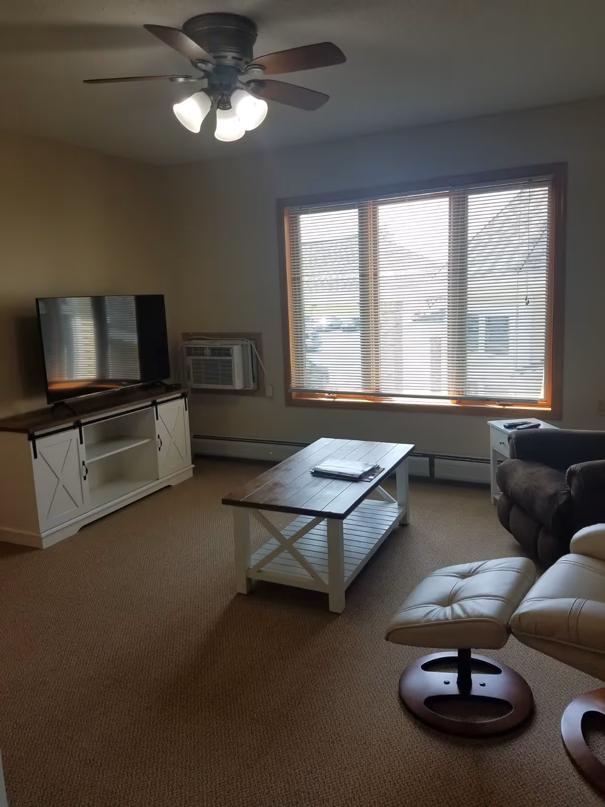 A living room with a ceiling fan light fixture, a large window with blinds, a flat-screen TV on a white wooden TV stand, a wooden coffee table with a magazine on it, a brown armchair, and a white leather recliner chair with a matching footrest.