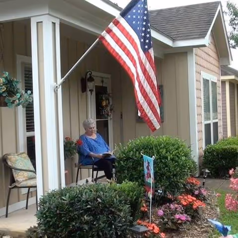 An elderly woman sitting on a chair on the porch of a beige house with white trim, reading a book. The porch is decorated with an American flag, potted plants, and a garden with colorful flowers and green bushes.