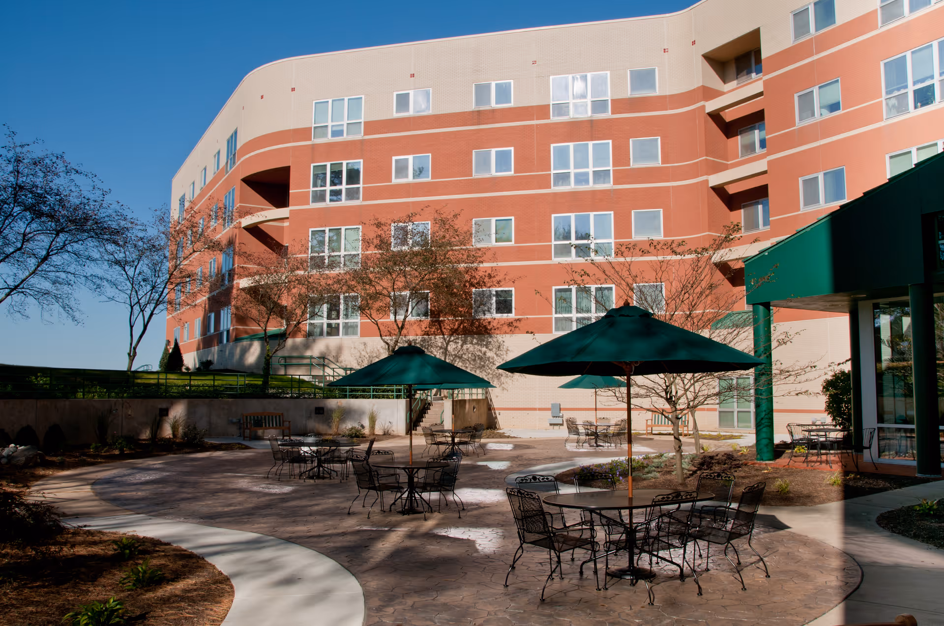 Outdoor patio area at Quarryville Presbyterian Retirement Community with several round metal tables and chairs, each shaded by green umbrellas. The patio is paved and surrounded by landscaped garden beds and trees, with a multi-story building in the background under a clear blue sky.