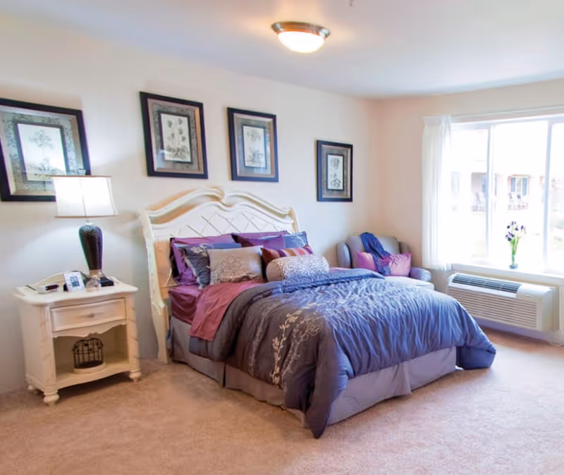 A cozy bedroom with a large bed dressed in purple and gray bedding, a white nightstand with a lamp and phone, four framed botanical prints on the wall above the bed, a cushioned armchair with pillows near a large window, and a wall-mounted air conditioning unit below the window.