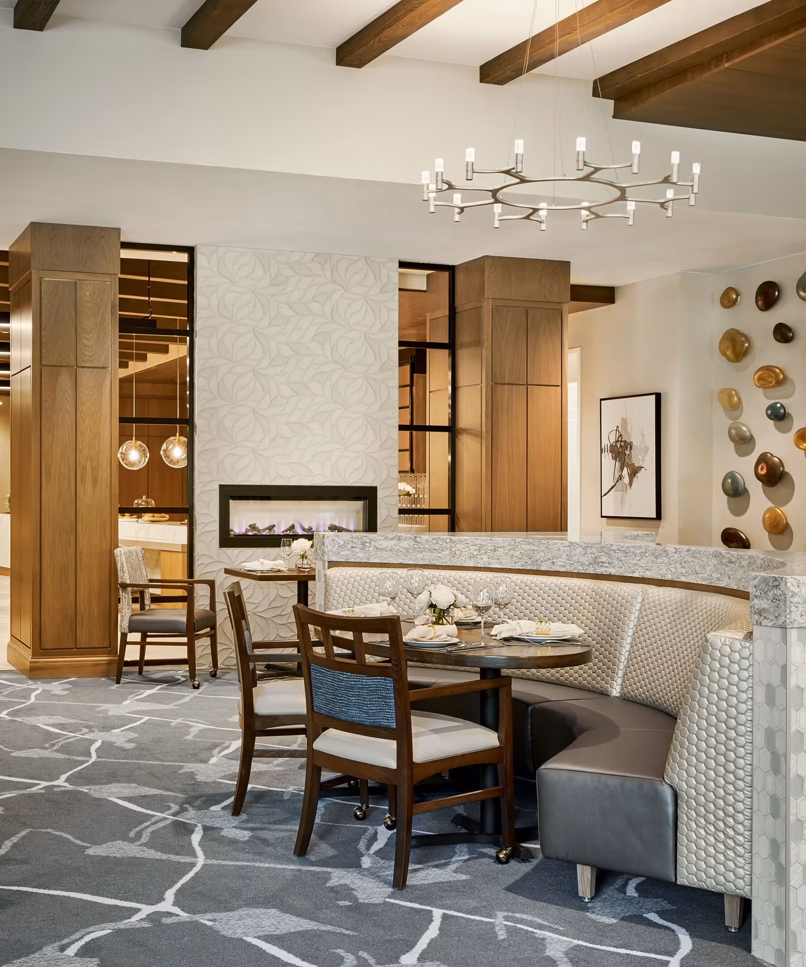A modern dining area with a curved booth upholstered in textured light-colored fabric and dark leather, accompanied by wooden chairs with light cushions. The room features a decorative fireplace with a leaf-patterned surround, wooden beams on the ceiling, a contemporary circular chandelier, and abstract wall art. The carpet has a gray and white abstract pattern.