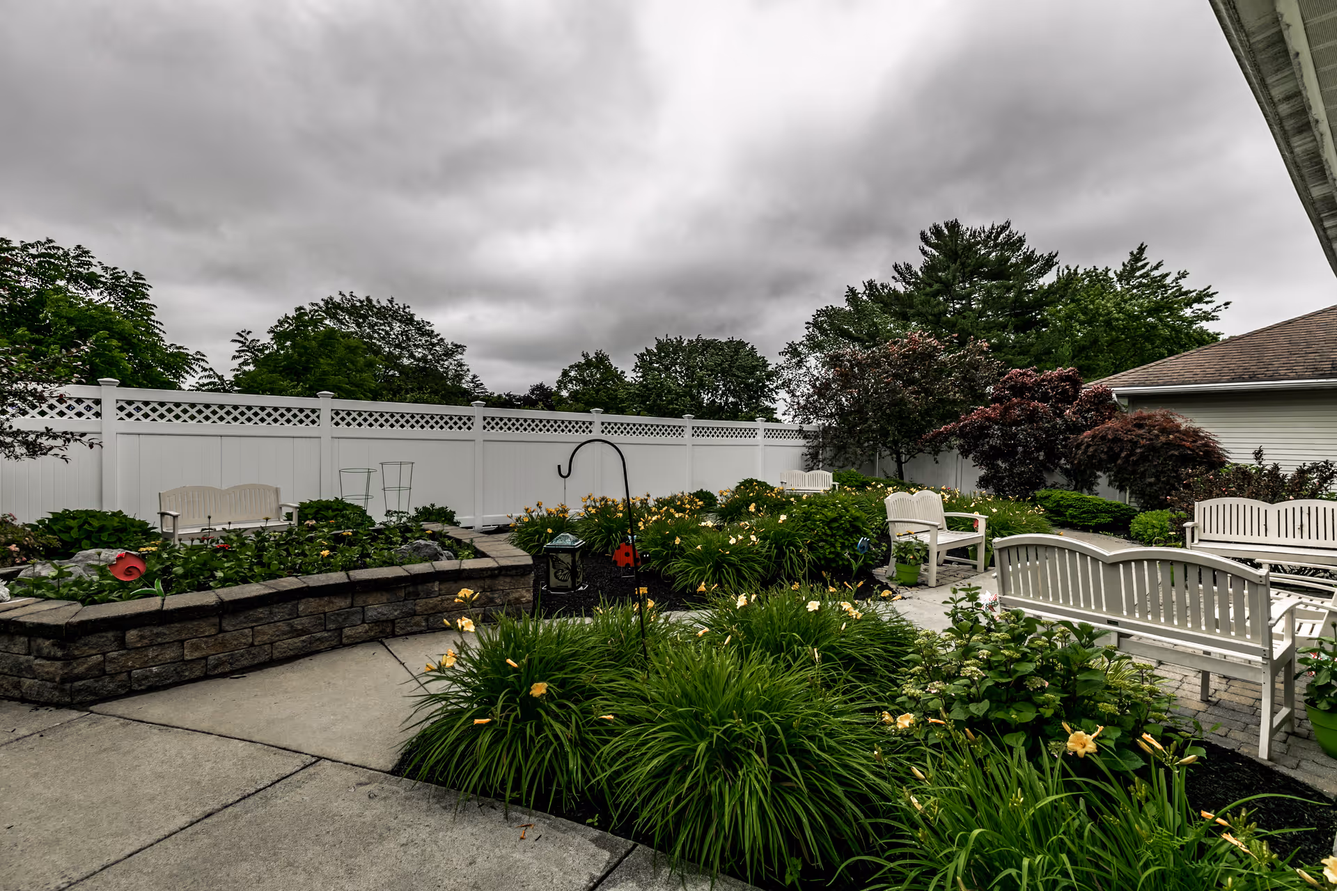 Outdoor garden area at Keepsake Village at Greenpoint with white benches, green shrubs, flowering plants, and a white fence under a cloudy sky.